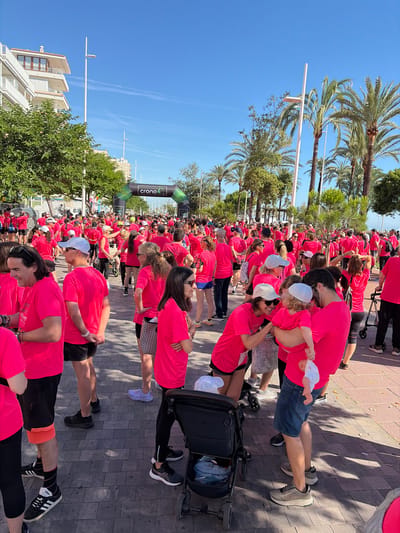 The image shows a large group of people, all wearing bright pink t-shirts, gathered for an event in Grau I Platja, Spain. They are standing on a paved walkway lined with palm trees, with buildings visible in the background. An archway with "Crono 4" is set up near the front of the crowd. Some individuals are carrying small children or pushing strollers. The day is sunny, casting shadows on the walkway.