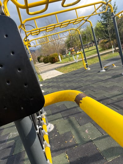This is a medium-shot, eye-level photograph taken in Chavdar, Bulgaria, of playground equipment. A black, textured, plastic seat with multiple small, circular indentations is prominently featured in the lower foreground. A grey metal pole runs horizontally behind the seat. To the left is a climbing frame with yellow metal rungs. A thick, yellow metal pipe with a patch of missing paint curves in the foreground. The playground surface is composed of dark green and grey rubber tiles. In the background, there are more playground structures, a grassy area, bare trees, bushes, and a light blue sky with clouds.