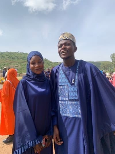 The image shows two people in what appears to be Naraguta Hausawa, Nigeria. One is a man wearing a blue traditional outfit with patterned embroidery, and a patterned hat. The other is a woman wearing a blue head covering and matching outfit. The setting appears to be outdoors, possibly at an event, with other people and some greenery in the background. The sky is overcast.