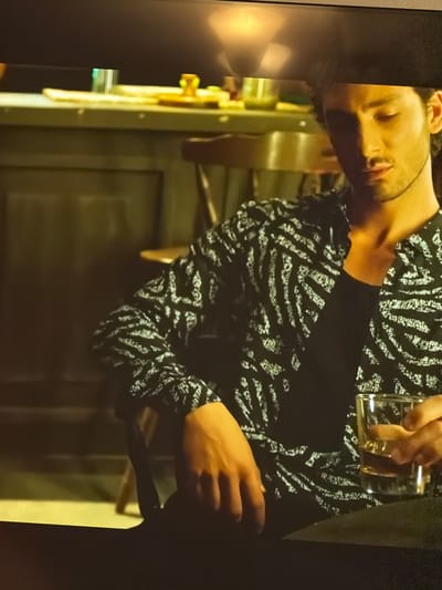 A man with olive skin and short, dark hair is visible in the image. He is wearing a black top with a white, zebra-like pattern. He is holding a glass of clear liquid, possibly alcohol. The background appears to be an interior setting, possibly a bar or restaurant, with a metallic structure or fixture visible on the left. The location is Bearpark, United Kingdom, but the information about the city is not obvious in the image. The man appears to be either drinking or having a drink poured for him.