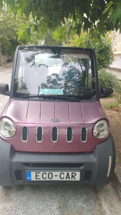 An outdoor, eye-level, front-facing shot captures a small, two-door, maroon colored electric car parked on a gravel surface. The vehicle has a black bumper with a European license plate displaying "GR ECO-CAR". The car's headlights are circular and prominent on either side of the front grille, which features five vertical, chrome-trimmed openings. Above the grille, a metallic logo is visible on the hood. The car's windshield reflects the surrounding environment, including trees with green leaves and a portion of the sky. The background shows foliage and hints of buildings, suggesting an urban or suburban setting. The car appears to be in good condition, parked at what is likely a roadside.