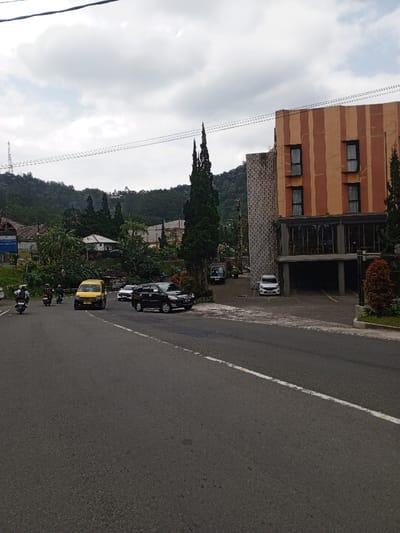 The scene depicts a roadside view in Ciloto, Indonesia, under an overcast sky. A multi-lane asphalt road with a solid white edge line and a faded dashed white lane line occupies the foreground and mid-ground.
On the left side of the road, two individuals are riding motorcycles, both wearing helmets. Behind them, a yellow van is stationary, facing the camera. Further back on the left, residential structures with light-colored roofs are visible amongst dense tropical vegetation. A communication tower is present on a distant green hillside.
On the right, a large, multi-story building with a distinctive facade features vertical orange and brown stripes, along with four prominent rectangular windows on its upper floors. An adjacent section of the building exhibits a light-colored, textured exterior. The ground floor of this building features large glass windows and an open, covered parking area. A black SUV is parked on an asphalt driveway directly off the main road, to the right of the yellow van. A white vehicle, possibly an MPV or SUV, is parked further back within the same property's parking area. A dark pickup truck is also visible near the entrance to this property. Tall, slender evergreen trees stand prominently in front of and around the building, with additional trees and bushes lining the road and properties.
The background consists of rolling, densely forested hills and mountains beneath a cloudy, grey sky, with power lines crossing horizontally near the top of the frame.