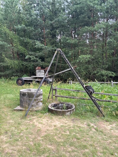 An outdoor scene in Sielachowskie, Poland, features a grassy clearing with a backdrop of dense evergreen trees. In the foreground, two circular concrete structures, likely old wells, are visible. To the left is a larger, taller well, with a wooden pallet beneath it. To the right is a smaller, shallower well. A rustic wooden fence runs horizontally across the mid-ground. A triangular metal structure, possibly a makeshift crane or derrick, stands prominently in the center, with a pulley mechanism attached to its upper section. Behind the wells and fence, a weathered wooden trailer is parked. It has two large wheels, one of which is partially obscured. On the trailer, a piece of rusty machinery with a wooden component is mounted. The overall impression is one of an abandoned or disused area, likely on the outskirts of the settlement.