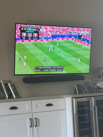 The image shows a large flat-screen television displaying a soccer match, likely from the FIFA World Cup, with "CAN 0" and "0 MAR" visible in the scoreline and "1:13" in the game time. A commentary panel with three commentators' photos and names—DEREK RAE, ROB GREEN, and DR. JOE MACHNIK—is present. Below the match, a caption reads, "AT THEIR DEFENSE, THE CENTER BACKS - A NEW PAIRING AGAIN." The television is mounted above a white entertainment unit with cabinets and drawers, and a soundbar is placed beneath the TV. Several framed photographs and a football are displayed on top of the entertainment unit. The overall scene suggests a domestic setting, possibly a living room or media room, in Muskegon, Michigan, United States of America.