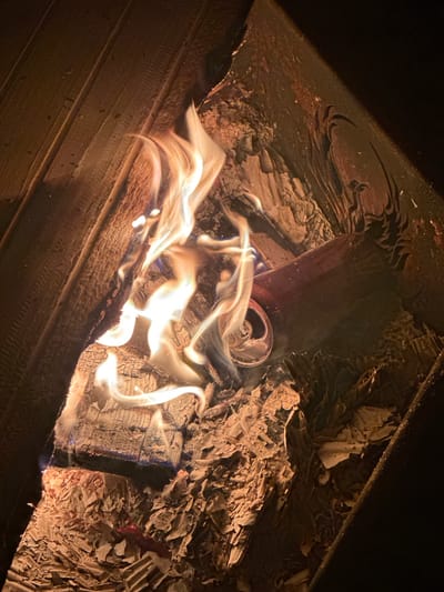 The image displays a close-up view of a burning fire, likely within a fire pit or fireplace, taken at night or in a dimly lit setting. The scene is dominated by bright, fluid flames in shades of yellow and orange, illuminating the surrounding materials.
The primary subjects are actively burning wood logs, characterized by their charred surfaces and glowing embers. Interspersed among the burning wood and ashes is a crumpled, partially melted aluminum can, resembling a soda can, which has been thrown into the fire. A substantial amount of ashes and smaller, partially consumed debris is spread around the burning materials, particularly on the right side of the frame.
The activity depicted is the active combustion of wood and other items in a fire. The flames are dynamic, with distinct wisps of light and heat rising from the burning mass. The environment enclosing the fire includes dark, possibly metal or heavily charred surfaces visible along the top and upper right, and distinct wooden planks forming a boundary or base in the lower-left section.
Notable details include the clear indication of nighttime due to the profound darkness surrounding the illuminated fire. The intense glow of the flames suggests significant heat. There are no visible people or discernible text within the image. Given the location context, this scene is set in Веревское, Russia. The image evokes a sense of warmth and the primal energy of fire.