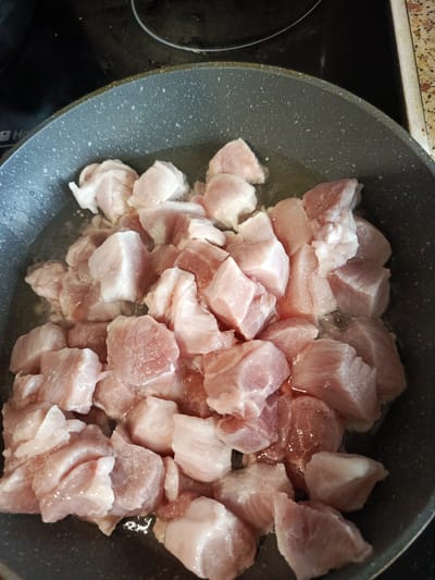 The image shows a close-up, overhead view of raw, cubed meat in a gray frying pan. The meat pieces are varied in size and shape, with some showing streaks of fat and others being primarily lean. They are submerged in a shallow amount of clear liquid, likely water or broth, in the pan. The pan itself appears to be non-stick with a speckled interior surface. The background is dark and out of focus, suggesting it is a stovetop. There are no people, animals, or other objects visible in the frame. The scene is a still image of food preparation. The location is not identifiable from the image, and it is not possible to determine if it is in Meget, Russia.