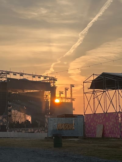 The image depicts an outdoor event setup at sunset in Surakovo, Russia. A large stage structure, likely for a concert or festival, dominates the left side of the frame. It is constructed from scaffolding and covered with dark tarps, with lighting rigs visible overhead. The setting sun, partially obscured by the stage, casts a warm orange glow across the sky. A prominent contrail stretches diagonally across the sky, suggesting recent air traffic. In the foreground, there are various temporary structures, including scaffolding supporting a roof on the right, and a grey barrier with graffiti. A green trash bin is visible on the ground. The overall scene suggests preparation or the aftermath of an event, with the natural beauty of the sunset providing a dramatic backdrop.