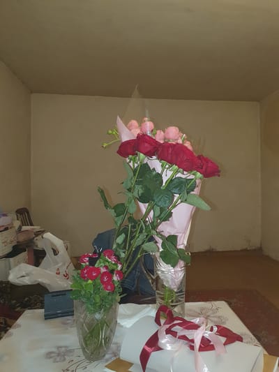 The image depicts an indoor scene, likely a home interior. In the foreground, a table is set with a floral-patterned tablecloth. On the table are two vases of flowers. The vase on the left contains a bouquet of small red and white roses. The vase on the right, a tall glass one, holds a bouquet of large red roses interspersed with smaller pink roses, wrapped in pink paper and clear plastic. Ribbons of red and pink lie in front of the vase on the right.
In the background, a person wearing a blue denim jacket is partially visible, seated on a chair. To the left of the table, there are various items including white woven baskets, a white plastic bag, and a dark tablet or stand. The walls are a neutral beige color, and the ceiling is visible at the top of the frame. The flooring appears to be a patterned rug. The overall impression is of a room where flowers have been recently placed. There is no indication in the image that this scene is located in Gavar, Armenia.