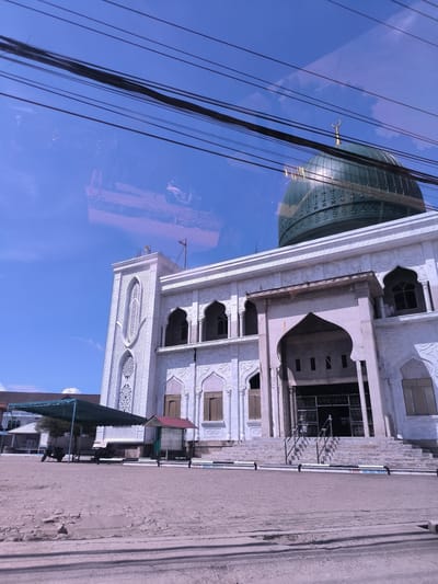 The image shows a large, white mosque with a prominent green dome, captured on a clear, sunny day. The photograph appears to be taken through a glass surface, as evidenced by reflections of overhead power lines, clouds, and faint shapes that might include a motorcycle and other elements of an outdoor scene, particularly visible in the upper and lower left portions of the image.

The mosque itself is a striking white building adorned with intricate geometric carvings and arched windows. A tall section on the left side of the mosque, possibly a minaret or a decorative tower, also features these detailed patterns. The main entrance is grand, with a large arched doorway leading to steps with metal railings. The green dome is topped with a golden crescent moon finial and features decorative bands.

In the foreground, there is an unpaved, dusty or gravelly area leading up to the mosque's steps. To the left of the mosque, a green canopy provides shade, and a small, red-roofed kiosk or shelter is also visible. Near the base of the mosque steps, low barriers are in place, one of which clearly displays the text "STOP SANDAL SEPATU" (Stop Sandals Shoes).

The weather is bright and sunny, indicated by the clear blue sky and strong shadows. No people are explicitly visible in the main scene. The image captures the mosque's architectural beauty within its immediate surroundings in Keudelapang, Indonesia.