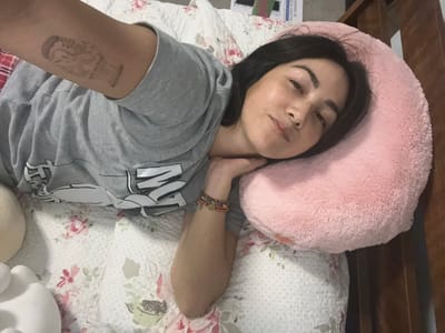 A woman is lying on a bed, with her head resting on a pink pillow. She is wearing a gray t-shirt and has a tattoo on her left arm. The bed has a floral patterned comforter. The location is Santiago de Surco, Peru.