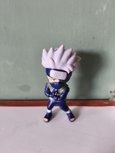 The image displays a miniature plastic figurine of the character Kakashi Hatake, from the "Naruto" series, standing upright and centered on a flat, light grey surface. The figurine exhibits white spiky hair, a dark blue forehead protector with a silver-colored Konoha symbol, a dark blue face mask, a dark blue uniform, a light green tactical vest, and dark blue open-toe sandals over white tabi socks. Its hands are clasped together at chest height, and a single right eye is visible above the mask. The immediate background features a horizontal reddish-brown wooden strip, above which is a faded, light greenish-grey wall showing minor discoloration. Soft illumination from the front or left casts a subtle shadow of the figurine to its right against the wall. This scene is situated in an indoor setting in Chimbote, Peru.