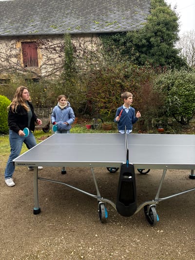 This vertically oriented image captures three individuals playing outdoor ping pong in a rustic garden or backyard setting.

In the foreground, a young woman with long, light-brown hair, wearing a black jacket and blue jeans, holds a blue ping pong paddle. She appears engaged in the game, positioned to strike the ball. To her right, across the ping pong table, a young boy with short, light-brown hair, dressed in a blue sweater over a collared shirt, holds a red paddle and looks upwards, seemingly following the trajectory of a ball. Further back and slightly to the left of the boy, another person, possibly a child, is visible, wearing a blue puffer jacket and a light-colored scarf, holding a blue ping pong paddle.

A prominent grey outdoor ping pong table with a black net occupies the center of the frame, separating the players. The brand "DECATHLON PONGORI" is visible in white text on the side of the table. The playing surface is slightly wet or reflective, possibly from recent rain or dew. The ground beneath the table and players is a light-colored gravel or dirt.

The background features lush green foliage from trees and bushes, suggesting a natural and unmanicured garden. An old, light-colored stone building with a tiled roof, partially covered in moss, is visible behind the greenery, along with a weathered wooden door. The sky is overcast, indicating a daytime scene, and the heavy clothing worn by the individuals suggests cool weather. The overall impression is one of casual outdoor recreation in a tranquil, rural setting, consistent with the provided location context of Étréchy, France.
