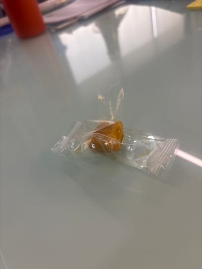 A piece of what appears to be a brown, cube-shaped food item encased in clear plastic packaging sits on a light-colored, reflective surface. The packaging is sealed at the top and bottom with crimped edges. Part of an orange and white object is visible in the lower left corner of the image. The environment is indistinct. The city of Buenavista, Mexico, is not discernible in the image.