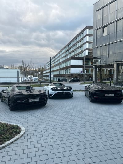 The image depicts an outdoor scene in Glattbrugg, Switzerland, showcasing three parked McLaren sports cars on a paved area adjacent to a modern office building. The building features a glass and steel facade, reflecting the cloudy sky. The cars, two black and one white, are parked in a row. Swiss license plates are visible on the front and rear of the vehicles. A small patch of greenery borders the paving. No people are visible in the frame.