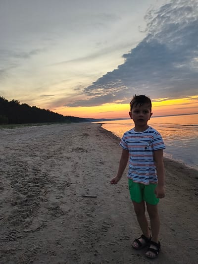A young boy stands on a sandy beach at dusk. He is wearing a striped t-shirt with red, blue, and white stripes, and bright green shorts. He is also wearing black sandals. The boy is looking directly at the camera with a neutral expression.

The setting is a wide, sandy beach that stretches into the distance. The sea is calm and reflects the colors of the sunset. A dark line of trees forms the backdrop to the beach, suggesting a forested area. The sky is filled with dramatic clouds and vibrant sunset colors, ranging from pale yellow and orange near the horizon to grayer tones higher up. The overall mood is serene and peaceful, typical of a sunset.

The time of day is clearly dusk, with the sun low on the horizon, casting a warm glow over the scene. The weather appears calm, with no strong winds indicated by the water or the boy's hair. The location is identified as Apšuciems, Latvia, which is known for its coastal landscapes and beaches.

There is a small, cartoonish character visible on the boy's t-shirt, appearing to be Snoopy. No other significant objects or text are visible in the image.