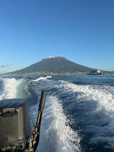 A wide-angle, eye-level shot depicts a view from a moving boat on a calm, blue sea. The boat's black outboard motor is visible in the foreground on the left. The water is disturbed, creating white frothy wakes that trail behind the boat. In the mid-ground, a large, green, cone-shaped volcano, Mount Kie Matubu, dominates the landscape, its peak partially obscured by a wispy white cloud. A coastal city, Tidore, with densely packed buildings in white and terracotta, sprawls across the base of the volcano. Further out at sea, a white and orange ferry boat is visible. The sky is a clear, bright blue with a few small clouds. The overall scene suggests a sunny day with tranquil seas, potentially in the early morning or late afternoon given the light. The location is identified as Tidore Islands, Indonesia.
