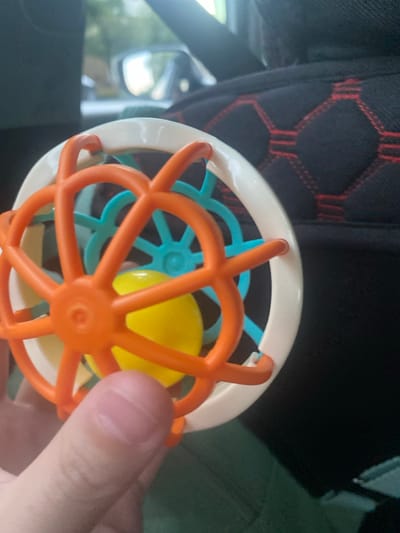 A hand is holding a colorful baby rattle. The rattle has an orange, white, and teal plastic structure with a yellow ball inside. The background is dark and appears to be the interior of a vehicle, possibly a car, with a black and red patterned fabric visible. There are some blurry reflections in the window of the vehicle, suggesting it is daytime. The overall scene appears to be casual and domestic. There is no visible text in the image.