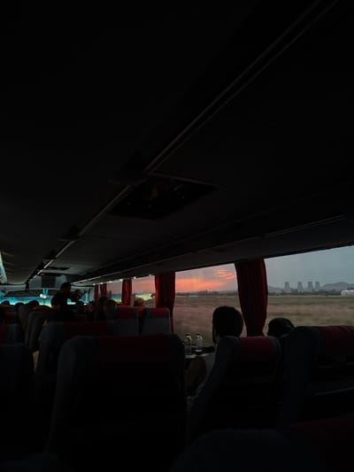 The image shows the interior of a bus traveling through the city of Arshaluys, Armenia, at the coordinates 40.157 latitude and 44.207 longitude. It is evening and the sun is setting outside, casting an orange glow on the sky and the landscape. There are several people sitting in the seats, most facing the back of the bus. There are two passengers, one in the front row and the other in the middle, with their backs towards the camera. The person in the front row has dark hair and is wearing a black jacket. The person in the middle row has dark hair and is wearing a white shirt. There are three more passengers seated in the back row, their faces are not visible. All of the passengers are facing the front of the bus, looking out the windows at the setting sun. The seats are mostly dark and the walls of the bus are also dark, making it difficult to see the passengers in the back row. The seats are dark grey and red, with a black leather covering. The windows are dark and there are red curtains on them. The bus is traveling along a road outside the city. There are industrial buildings in the distance to the right of the bus. The landscape is a mix of green and brown, with a dusty atmosphere. The image was taken from the perspective of a passenger seated in the front row looking toward the back of the bus.