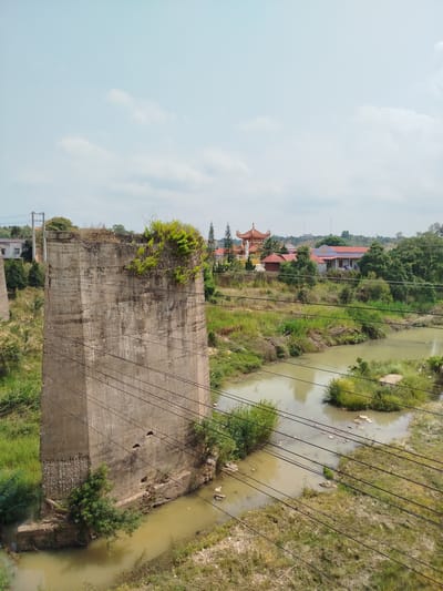 A tall, concrete pylon stands in the foreground, topped with vegetation, situated within a river landscape in Đại Ninh, Vietnam. The river, reflecting a muddy color, weaves between grassy banks and small islands of vegetation. Power lines traverse the scene diagonally from top to bottom. In the mid-ground, a complex of buildings with red-tiled roofs, including what appears to be a temple structure, emerges from a dense cluster of trees and greenery. The sky is overcast with a pale blue hue, lightly dotted with clouds.