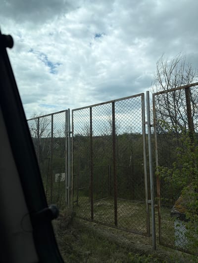The image presents a view through a chain-link fence. Beyond the fence, a vegetation-covered area with trees and grass is visible. The sky is overcast with cloudy conditions. The image is rotated, suggesting the perspective is from within a vehicle. The specific location in Dulevo, Bulgaria, cannot be determined from the visual information provided. No people or actions are visible.