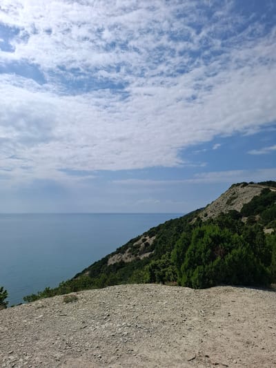 The image shows a coastal landscape with a steep, rocky cliff covered in sparse vegetation and evergreen trees, overlooking a calm, blue sea. The sky is partly cloudy with white, fluffy clouds against a bright blue background. The foreground is a rocky, gravelly surface. There are no people or animals visible in the image. The scene appears to be a natural, undeveloped coastal area. The location is identified as the city of Varvarovka, Russia.