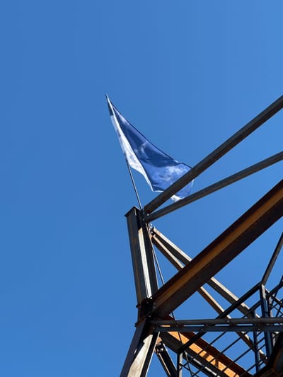 A blue and white flag is waving from the top of a metal structure against a clear blue sky. The metal structure appears to be part of an industrial or construction site, with rusted beams and supports visible. The perspective is from below, looking upwards at the flag and the sky. The scene does not provide any specific visual cues to identify the location as Yelets, Russia.