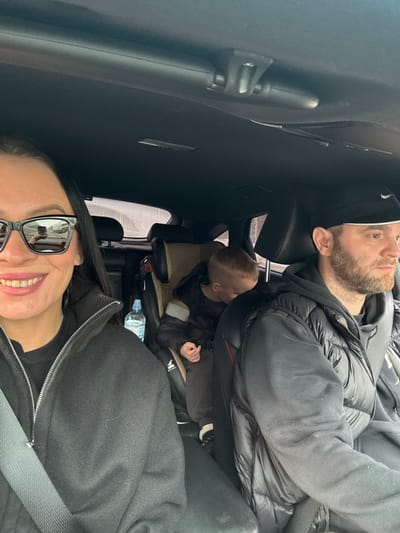Within a vehicle in Kabanov Mys, Russia, three individuals are present. A female is positioned in the front-left seat, wearing black sunglasses that reflect a road scene, a dark zippered jacket, and a visible seatbelt; she is smiling at the camera. A clear plastic bottle with a blue cap rests to her right. In the front-right seat, a bearded male, wearing a black baseball cap and a dark puffy vest, is observed looking forward, indicating operation of the vehicle, with a seatbelt visible across his chest. A light-haired child is secured in a tan child safety seat in the rear-left, wearing a dark puffy coat, with its head bent forward. The vehicle's interior is uniformly dark, and external visibility through the windows is diffused.