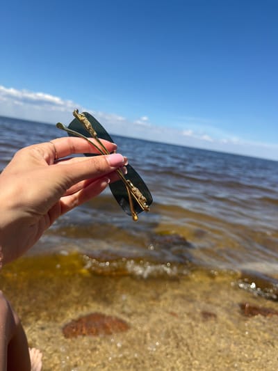 A person's hand is holding a pair of dark-lensed sunglasses with gold frames and ornate detailing. The background features a body of water, likely the Kyiv Reservoir near Vyshhorod, Ukraine, under a clear blue sky with sparse clouds. The water appears murky brown and shallow near the sandy shore, with visible rocks beneath the surface. The overall scene suggests a bright, sunny day at the water's edge.
