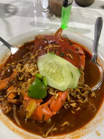 A high-angle forensic shot captures a dining scene in Alue Mudem, Indonesia, focused on a white ceramic bowl containing a main dish. The bowl holds instant-style noodles in a thick, dark reddish-brown sauce or broth, accompanied by a cooked crab with an intact, orange-red shell. Garnish includes two slices of green cucumber, a lime wedge, and two small green chili peppers. A silver-colored fork is visible on the left side of the bowl, and a matching spoon on the right. The bowl's rim shows traces of the sauce. In the blurred background on a white surface, a clear glass mug containing a light brown beverage is partially visible, positioned behind a neon green disposable lighter standing upright. A dark cylindrical object is partially discernible behind the mug. No individuals are present in the frame.