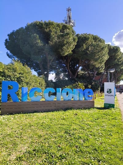 The image displays an outdoor scene in Riccione, Italy, centered on a large civic sign. In the foreground, green grass, punctuated by sparse white flowers and brown patches, covers the ground.
The primary subject is a multi-colored blue sign spelling "RICCIONE" in large, three-dimensional letters. The letters are painted in varying shades of blue, exhibiting a gradient or patterned effect, and rest on a light brown, slatted base positioned horizontally. This sign is situated in the lower-mid section of the frame.
To the immediate right of the "RICCIONE" sign stands a tall, rectangular white banner or signpost. This banner features green segments at its top and bottom. The top green section contains a crest-like logo and text including "1933 RICcione," while below, the phrase "GENERAZIONE S" is written in black. Additional smaller logos, such as "BSG" and "PUMA," are visible near the bottom of the banner.
Behind the signage, a dense array of green foliage, including trees and bushes, forms a natural background. Dominating the upper background is a prominent row of large, mature pine trees with distinctive broad, flat canopies. Above these trees, particularly on the upper right, a metallic cellular communication tower with multiple antenna arrays is clearly visible against a bright, clear blue sky. A street light fixture with two luminaires is mounted on a pole visible to the right of the banner, partially obscured by the trees.
In the far right background, beyond the banner and trees, a paved pathway or road is discernible. Two indistinct human figures are observed walking from left to right on this path. The sky is predominantly clear blue, with a small cloud formation visible on the far right edge of the frame. The scene is illuminated by direct sunlight.