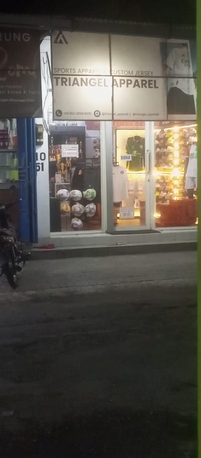 The image shows the storefront of "Triangle Apparel" located at -7.35 latitude and 112.764 longitude in Kecamatan Waru, Indonesia. There are three glass display windows, one of which is partially obscured by a blue pillar. The shop sells sports apparel and custom jerseys. The storefront sign is white with a black triangle and the words "Sports Apparel," "Custom Jersey," and "Triangle Apparel," along with contact information. There are various items on display inside the shop, including several soccer balls, a white shirt with a design, a green soccer jersey, and several pairs of shoes. The "Buka" sign indicates the shop is open. There is a motorcycle parked in front of the shop. The sidewalk is made of concrete pavers. The image appears to be taken at night.