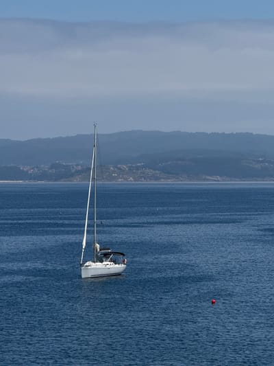 A white sailboat is anchored in the blue ocean near the island of Ons, Spain. The boat has a single mast and is partially covered by a dark canopy. The island of Ons is visible in the background as a hazy, green landmass with some buildings scattered across it. The sky is a pale blue with some light clouds. There is a red buoy floating in the water to the right of the sailboat. The water is calm with gentle ripples.
