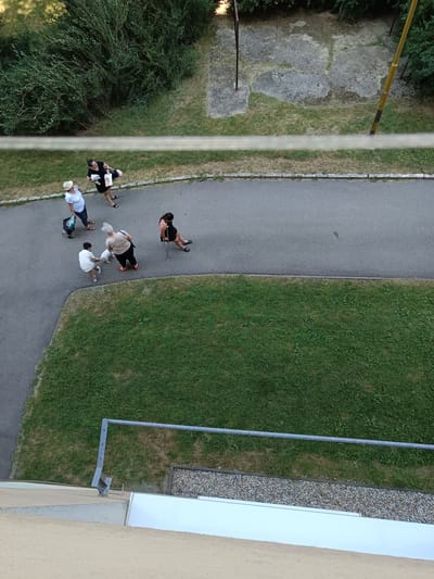 The image, taken from a high vantage point, depicts a sidewalk adjacent to a grassy area in the city of Košice, Slovakia. Several individuals are present. In the upper portion of the image, two women are walking. One is wearing a white hat and shirt, and the other a black dress. Further down the sidewalk, a woman with a white-haired ponytail is walking a small white dog. Next to her, a child in a white outfit is interacting with the dog. To the right, a woman with dark hair is walking with the aid of a crutch. The environment includes dense green foliage, a paved walkway, a lawn, and a concrete area with some debris. A metal railing is visible in the lower portion of the image.