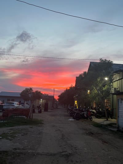 The image depicts a street scene in Keudekruenggeukueh, Indonesia, at dusk. The sky is a vibrant gradient of pink and orange, transitioning to pale blue, with scattered clouds. On the left side of the street, several cars are parked in front of buildings. A stack of terracotta tiles is visible on the ground. The right side of the street features buildings with outdoor lighting and several motorcycles parked along the roadside. The ground is unpaved and appears to be a dirt road. No people are clearly visible in the image. The overall atmosphere is serene, with the dramatic sunset being the dominant feature.