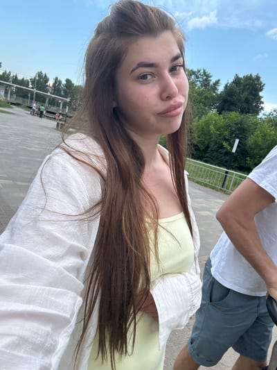 A young woman with long brown hair is taking a selfie outdoors. She is wearing a white open shirt over a light yellow top. Her hair is styled in a high ponytail. Her lips are slightly parted. Behind her, a man in a white t-shirt and grey shorts is partially visible to the right. The background shows a paved walkway, a metal fence, trees, and a cloudy blue sky. In the distance, there is a covered structure and several people. The location appears to be a park or recreational area in the city of Rechitsa, Russia.