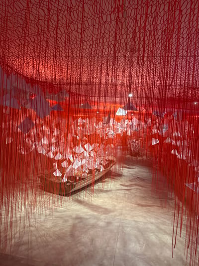 This image depicts an indoor art installation, stated to be located in San Isidro Llano Grande, Mexico. The scene is dominated by a dense, immersive environment of vertically suspended thin red threads extending from an overhead red, irregular netting structure. Numerous translucent, irregularly shaped white objects are interspersed among these threads, concentrated in the mid-ground, appearing illuminated. A dark-brown, narrow wooden boat, consistent with a canoe or traditional small vessel, rests on the light-colored, polished concrete floor in the mid-left. The space is illuminated by overhead artificial light sources, casting a strong red hue across the entire installation and generating discernible shadows on the floor. No individuals are visible within the frame.