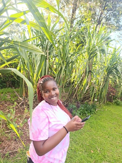 The image depicts a young woman standing in a grassy area near a patch of tall green foliage, possibly a sugar cane plantation. The woman is wearing a pink tie-dye t-shirt and has long reddish hair. She is holding a black smartphone in her right hand, with her left hand wrapped around her wrist. Her smile suggests she is interacting with the device. The background features lush greenery, with the sky visible through the foliage. The location of the image, based on the provided coordinates, is Kaplelwo, Kenya. The scene is characterized by a relaxed and casual atmosphere. The woman's interaction with the smartphone suggests a personal or social activity, while the lush greenery suggests a rural setting. The image captures a moment of everyday life in Kaplelwo, Kenya.