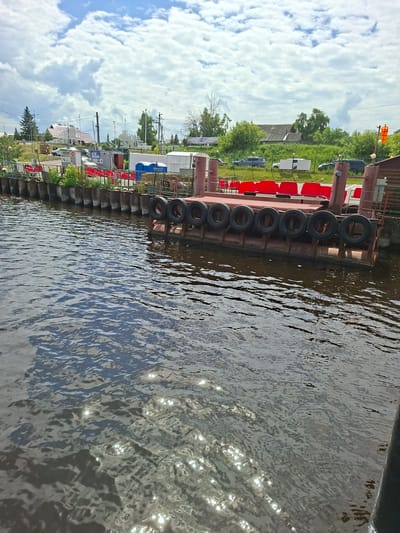 This is a medium-angle, eye-level shot of a ferry landing at the city of Verkhny Uslon, Russia. The ferry landing is made of metal and is lined with tires, which serve as bumpers. Red seats are visible on the ferry, and there are several structures behind the landing, including buildings and trees. The water in the foreground is rippled and reflects the sunlight. The sky is cloudy, with patches of blue visible. The image is taken on a sunny day.