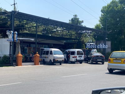 This is a street-level, eye-level, medium shot of a "Molochka" (dairy product store) in Andijan City, Uzbekistan, on a bright, sunny day. A black Chevrolet sedan is parked in front of the store, with its driver's side door open. Next to it, two white minivans are parked. The left minivan has a license plate that reads "T 512 FB". The right minivan has a license plate that reads "60 H009 MB". In the foreground, the rear of a yellow Daewoo car is visible, with the license plate "60 G 863 LA". The "Molochka" store has a large, dark metal awning supported by metal poles. A sign with the "Molochka" logo, featuring a cow, is visible on the left side of the store. The street is paved and appears to be in good condition, with some cracks. Trees and utility lines are visible in the background. The overall scene depicts a commercial area with vehicles and pedestrians.
