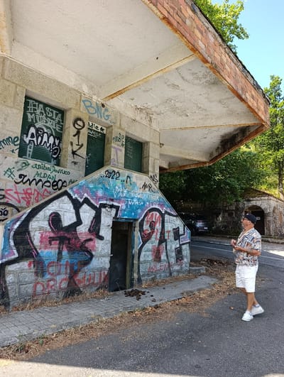 A man stands on a paved area next to a building covered in graffiti. The building appears abandoned, with a decaying roof supported by rusty beams and crumbling concrete. The man is wearing a patterned short-sleeved shirt, white shorts, a baseball cap, and white sneakers. He is looking upwards towards the building's roof, holding a small object in his right hand. The building has a staircase leading up to an entrance, also adorned with colorful graffiti. Beyond the building, trees and a road are visible. The ground is a mix of pavement and dry grass and leaves. The scene appears to be in a rural or semi-urban setting, possibly a derelict structure in Água da Pala, Portugal.