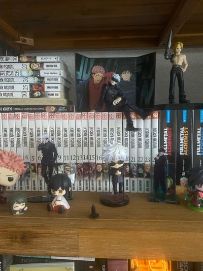 The image shows a wooden shelf filled with manga volumes and anime figurines. On the left side, there is a stack of Jujutsu Kaisen manga volumes. Further to the right, there are more Jujutsu Kaisen volumes and some Fullmetal Alchemist manga. Several anime figurines are displayed on the shelf, including characters from Jujutsu Kaisen and other series. One figurine of a male character with blonde hair and an arm blade stands on the top right of the shelf. In the middle of the shelf, a character with white hair and sunglasses is depicted, seemingly sitting. The background includes a poster or backdrop featuring characters from Jujutsu Kaisen. The lighting suggests an indoor environment. The image does not contain any information to determine its location in Hildesheim, Germany.