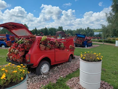 The image shows a sunny day in Kovalivka, Ukraine, with a clear blue sky dotted with white clouds. In the foreground, a bright red car is repurposed as a planter, with its hood and trunk open, overflowing with various potted flowers in shades of red, pink, and white. The car is situated on a bed of crushed red and grey gravel. To the right, a white cylindrical planter barrel also filled with yellow flowers stands in the grass. In the background, another blue car serves a similar purpose as a flower planter. Beyond the cars, a line of trees, a small wooden building, and a body of water can be seen. The overall scene suggests an outdoor decorative display in a park or garden setting. There are no people visible in the image.