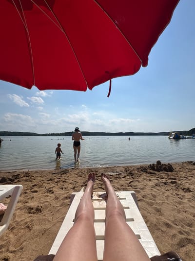 A person's legs are stretched out on a white lounge chair on a sandy beach under a large red umbrella. The beach is adjacent to a body of water, likely a lake, where other people are visible. In the water, a child stands near an adult who is facing away from the camera. Further out, there are several other people swimming and an inflatable water structure. The background shows a tree-lined horizon under a bright blue sky with scattered clouds. The scene appears to be a recreation area in the city of Ozyaritska-Slabadski rural council, Belarus.