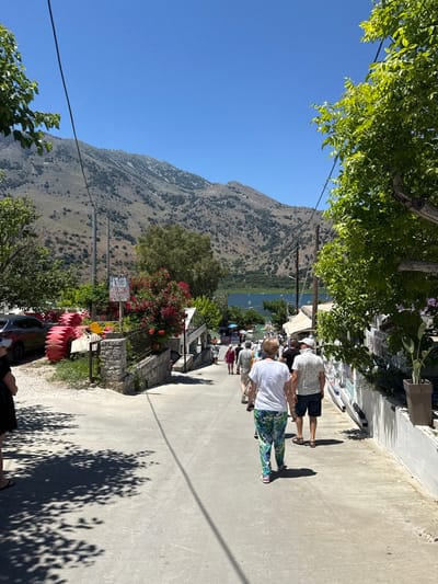 The image shows a street sloping down towards a body of water with mountains in the background, likely in Georgioupoli, Greece. Several people are walking down the street, some in casual summer clothing. There are trees and bushes lining the street, and some buildings and shops are visible. In the foreground, there are shadows of trees on the pavement. On the left side of the street, there is a car and some red objects that appear to be inflatable rafts. A signpost with red lettering is visible among some flowering bushes. The sky is clear and blue, indicating a sunny day. The scene appears to be a tourist area.