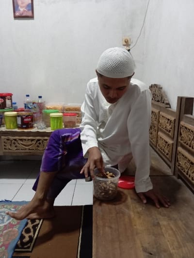 An adult male, wearing a white embroidered long-sleeved shirt and a white knitted kufi cap, is depicted indoors in the city of Bulukandang, Indonesia. He is in a crouching posture on a light wooden surface, with his right hand reaching into a clear cylindrical plastic container filled with light brown, irregularly shaped snacks. A red plastic lid is beside the container. His left hand rests flat on the wooden surface. He wears purple, patterned sarong-like trousers, and his bare left foot is visible on a patterned rug laid over a white tiled floor. To the left of the male, a wooden table holds an assortment of colorful plastic containers (red, green, yellow, brown) and plastic bottles of water. A carved wooden panel, possibly part of a bench or room divider, is visible to the right. The background consists of a plain white wall, featuring a framed picture of a baby or young child in the upper left corner and an electrical outlet or switch with visible wiring in the upper right.