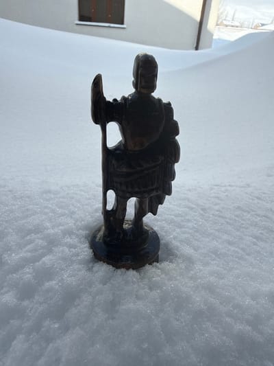 A dark, intricately carved figure stands upright in the snow. It appears to be a man carrying something on his back. The figure has a distinct head and legs with small openings where the legs connect to the body. A round, wide base supports the figure, partially buried in the snow. The surrounding area is covered in a thick layer of fresh snow with a building visible in the blurred background.