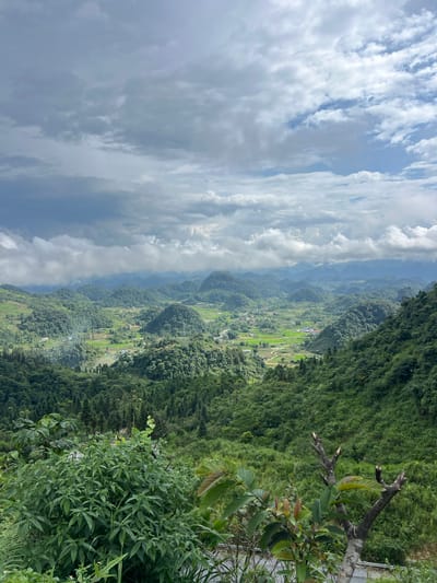 This is a high-angle, wide shot of a lush, mountainous landscape in Quản Bạ Commune, Vietnam. The foreground features dense green foliage with a bare, pruned tree trunk. Beyond this, rolling hills covered in trees and vegetation stretch into the distance. In the mid-ground, a valley opens up with a winding river or stream, interspersed with agricultural fields and small clusters of buildings, suggesting a village or hamlet. The sky above is dramatic, filled with a mix of dark, brooding storm clouds and brighter, sunlit clouds, casting a varied light across the scene. The overall impression is one of natural beauty and a rural, mountainous environment. There are no people or animals visible in the image.
