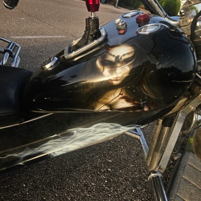 This is a close-up, eye-level shot of the fuel tank of a black motorcycle parked on an asphalt road. The motorcycle's fuel tank features a custom airbrushed design of a woman's face with dark hair, a jeweled headdress, and possibly blood splatters on her face and neck. The design is reflective and distorted due to the curved surface of the tank. White smoky graphics are also visible on the lower part of the tank.
In the background, the asphalt road surface is visible, marked with a faint white line. To the left and top of the frame, chrome handlebars and a red gear shifter with a black fabric boot are visible. The surrounding environment appears to be outdoors, with some greenery visible in the upper right corner. The image captures a detail of the motorcycle, focusing on its decorative elements and components. There are no people or actions depicted in the image. The location is the city of Illescas, Spain.