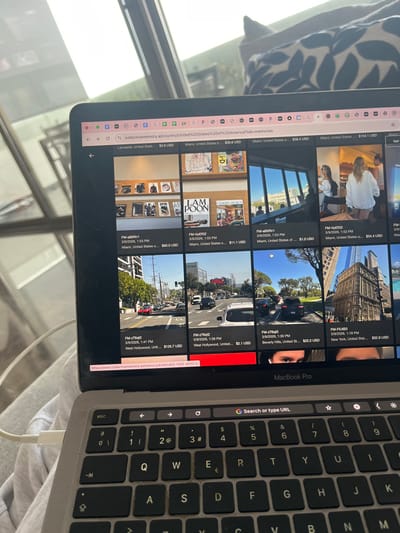 A MacBook Pro laptop is depicted at a slight angle, with its screen displaying a web browser interface. The laptop rests on a light grey, soft textile surface, likely clothing or a blanket. In the blurred background, natural light enters through a large window or glass door, revealing abstract reflections and hints of outdoor greenery and modern architectural structures. A dark cushion with a lighter, abstract pattern is partially visible in the upper right.
The laptop screen shows a Google Chrome browser window, with the URL `collectivememory.ai/country/United%20States%20of%20America?tab=memories` visible in the address bar. The screen displays a grid of thumbnail images from the website, each associated with a price in USD and a specific U.S. location (e.g., Lancaster, Miami, West Hollywood, Beverly Hills, New York). The images depict diverse subjects, including art displayed in interior spaces, busy city streets with vehicles, and exterior building facades. A white USB-C charging cable is plugged into the left side of the laptop. The laptop's keyboard, featuring a QWERTY layout with dark keys and white lettering, is visible in the lower portion of the image. "MacBook Pro" is printed at the bottom edge of the screen. No individuals are directly visible; however, the active laptop suggests a user is interacting with it. The image itself provides no visual information to place the scene in Santiago Yancuitlalpan, Mexico.