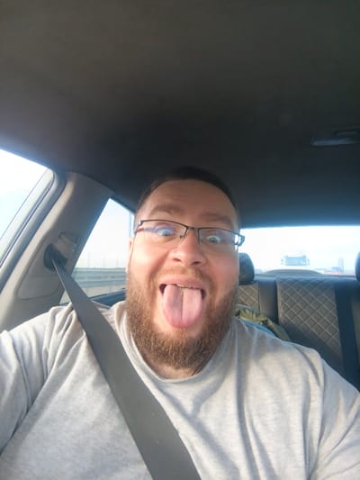 A man with a beard and glasses sticks his tongue out while sitting in the passenger seat of a car. He is wearing a gray t-shirt and a seatbelt. The car is on a road with other vehicles in the distance. The background appears to be a bright, possibly overcast sky. The scene is captured from a selfie perspective. The location is identified as Novosekki, Russia, suggesting it is a highway or suburban road.